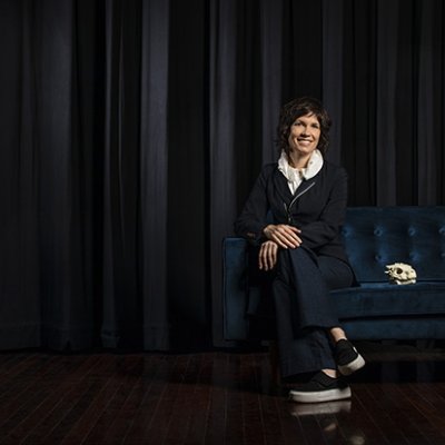 Picture of Deborah Riley sitting right of screen with black curtain behind her 